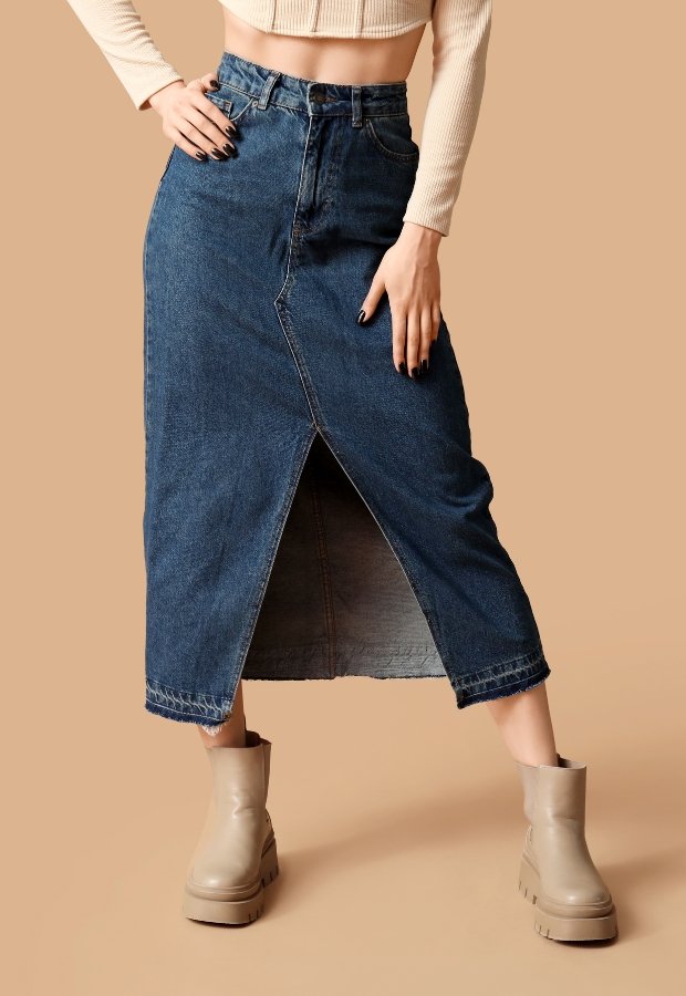 A model wearing a dark indigo denim A-line skirt with a front zipper, paired with a cropped beige knit sweater and chunky beige ankle boots. The skirt features a front slit and visible topstitching against a warm tan background. Denim A-Line Skirt
