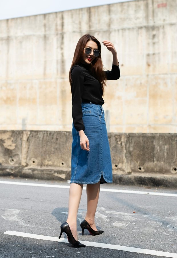 A stylish woman walks on a paved street wearing a black long-sleeve top, a knee-length blue denim A-line skirt, black high heels, and aviator sunglasses. She has long brown hair and red lipstick, posing with one hand brushing her hair against a light, textured wall backdrop.