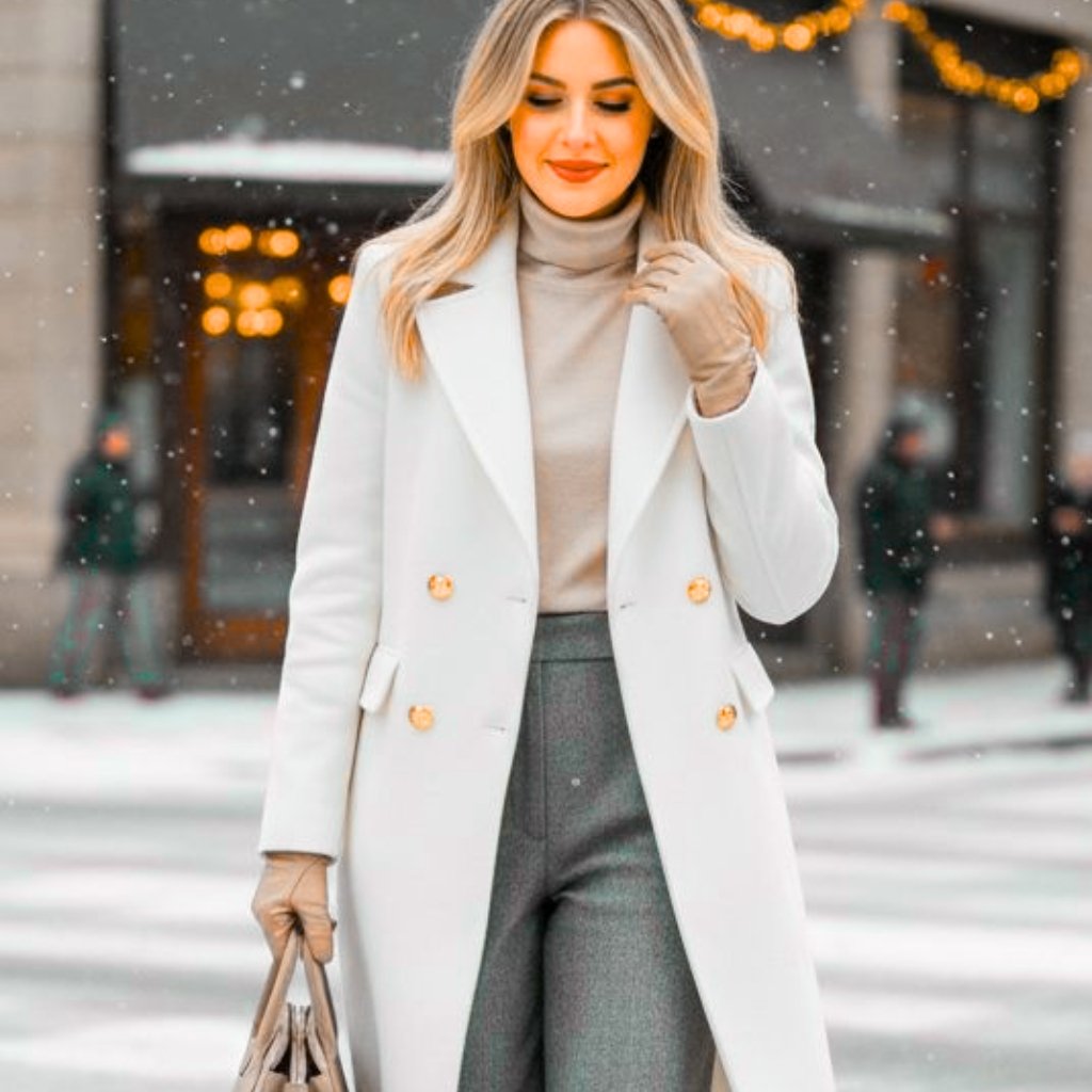 A stylish woman walking in a snowy urban scene, wearing a long white coat, beige turtleneck, gray tailored trousers, beige gloves, and carrying a beige handbag.
