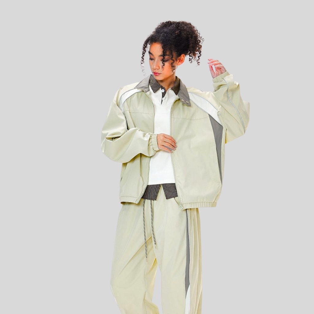 A person wearing a light beige oversized windbreaker and matching track pants with gray and white panels, standing against a light gray background. They have curly hair pulled back and are looking down while adjusting the sleeve.