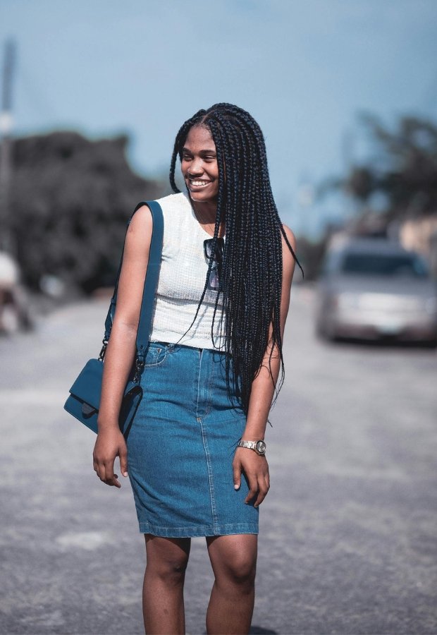 A smiling young woman with long braided hair wearing a white sleeveless top and a blue denim A-line skirt, accessorized with a blue crossbody bag and a watch, standing on a sunny street.