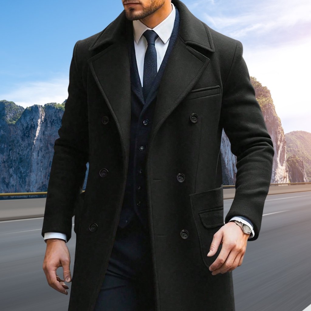A stylish man wearing a long black overcoat over a dark suit and white shirt, walking along a roadway with rocky cliffs and blue sky in the background.
