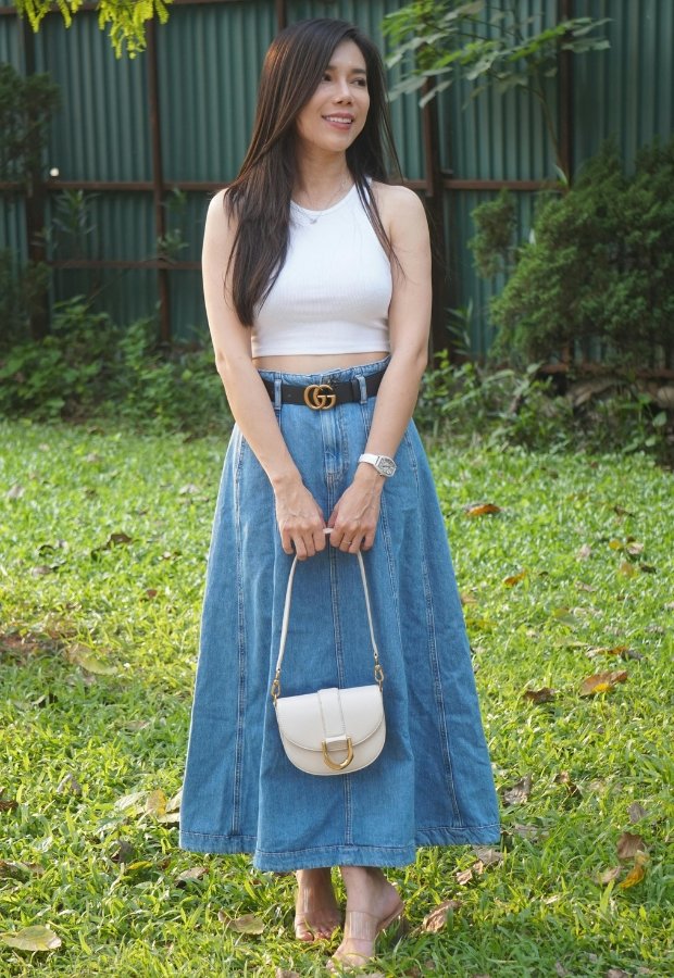 A stylish woman stands outdoors on a grassy lawn, wearing a white sleeveless crop top, a long high-waisted A-line denim skirt, a black Gucci belt, a small white handbag, and sandals. She smiles softly and looks off to the side.