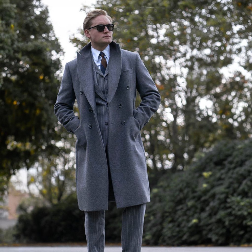 A man wearing a smart, double-breasted grey overcoat, grey suit, and sunglasses stands outdoors with hands in pockets.
