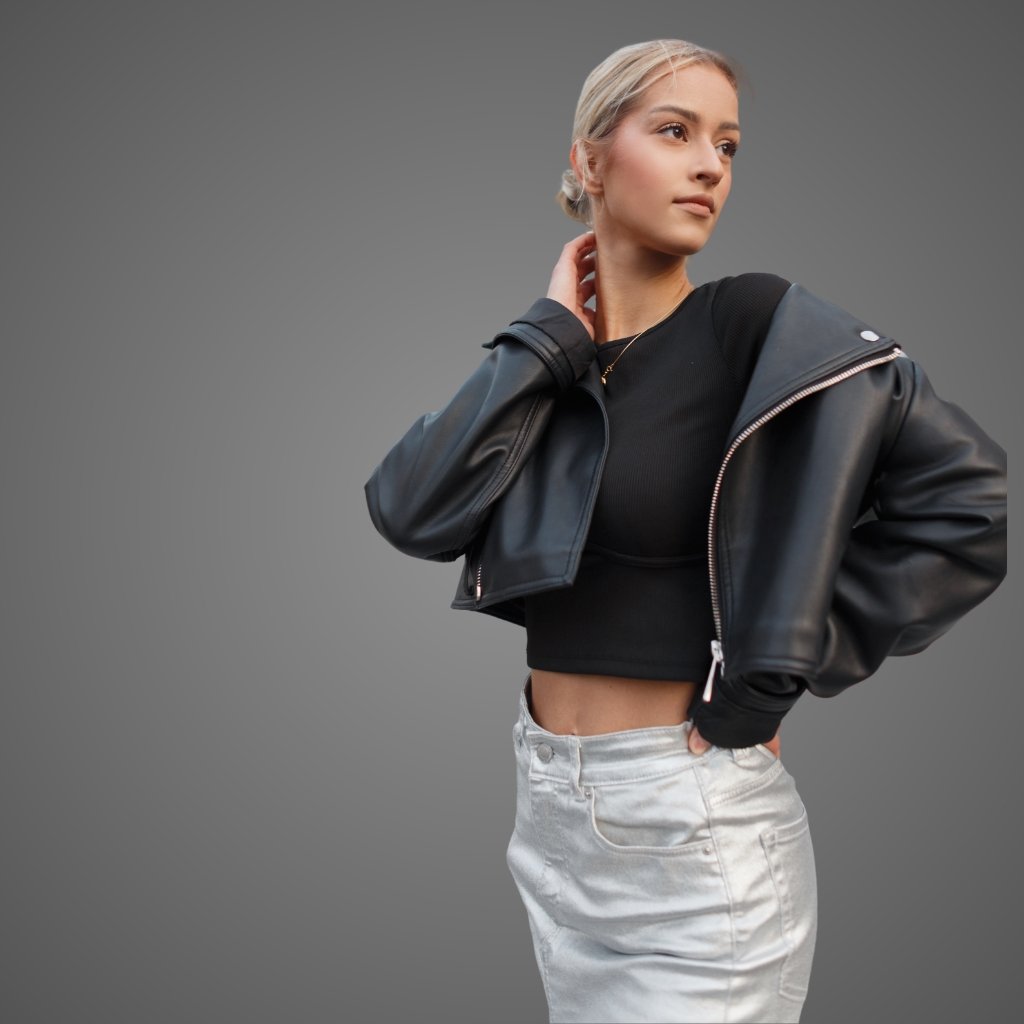 A stylish woman wearing a cropped black leather jacket over a black crop top, paired with light-wash jeans. Her blonde hair is slicked back, and she poses with one hand at her neck against a neutral gray background.Leather Short Jacket for Women