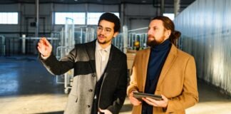 Topcoat History: How Classic Outerwear Evolved Through Time. Two men in a warehouse or industrial setting; one wearing a gray overcoat and glasses points toward something, while the other in a tan coat holds a tablet and looks on. Topcoat History