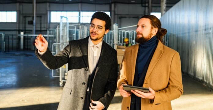 Two men in a warehouse or industrial setting; one wearing a gray overcoat and glasses points toward something, while the other in a tan coat holds a tablet and looks on. Topcoat History