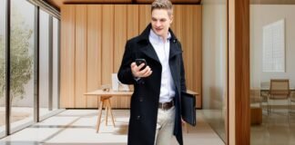 Topcoat Jackets for Men: Perfect Blend of Style & Warmth A man wearing a dark overcoat, light shirt, and beige trousers stands in a modern, sunlit office lobby, looking at his smartphone while holding a dark tablet case under his arm. Topcoat Jackets for Men