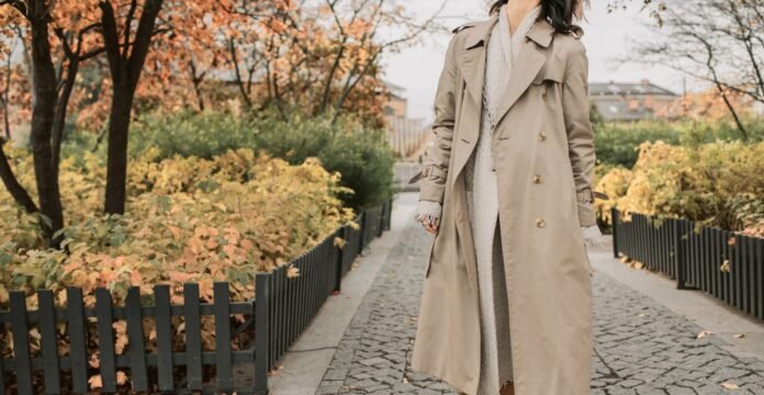 A person wearing a long beige trench coat over a light knit sweater and dress walks along a cobblestone path lined with autumn foliage and low black fences. Topcoat vs Overcoat