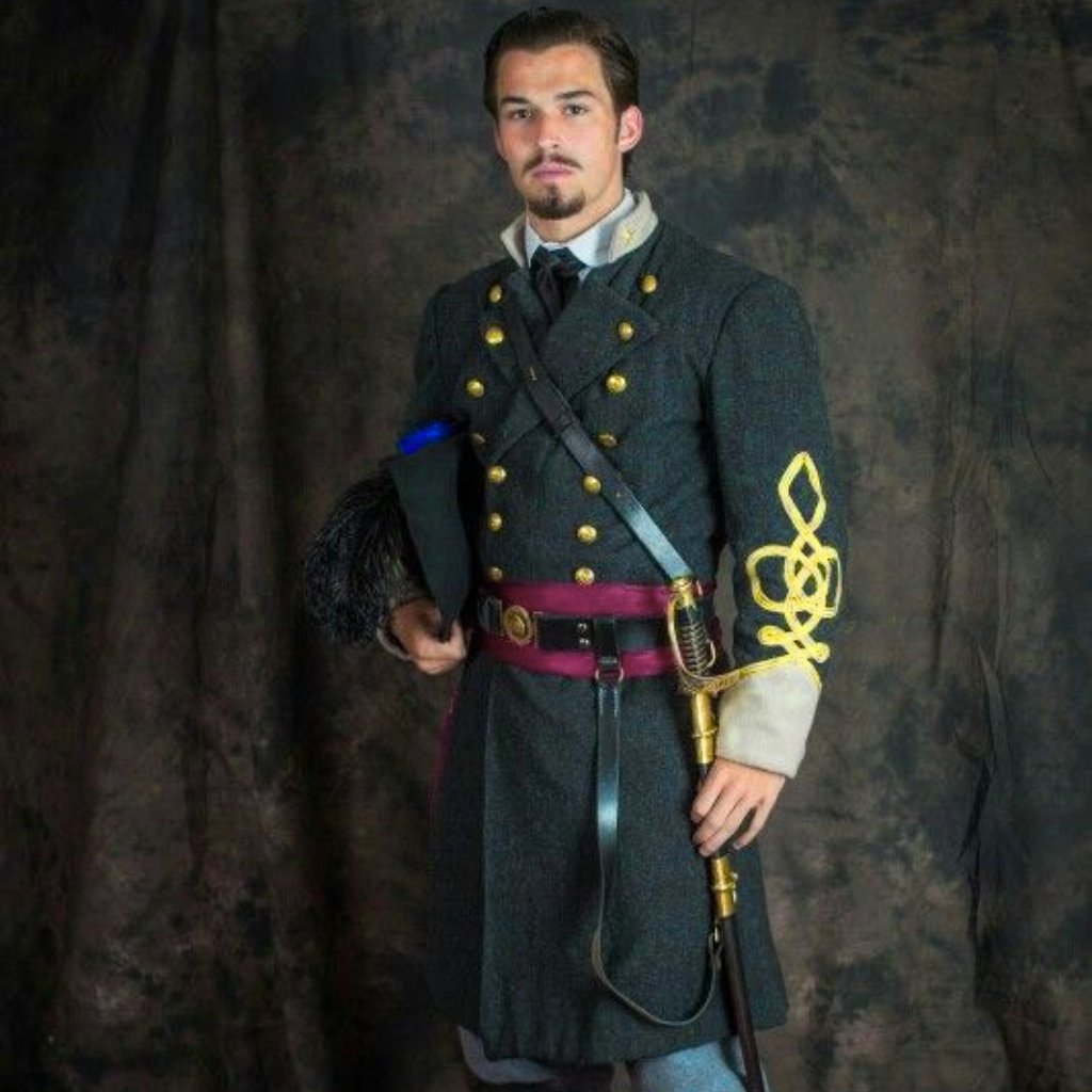 A man dressed in a dark military-style uniform with gold buttons, a magenta belt, and a decorative gold embroidered sleeve. He holds a black hat with a feather and a sword, standing against a dark backdrop.