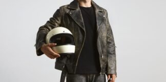 Travel Field Leather Jacket: Perfect Companion Every Trip A person wearing a weathered gray leather motorcycle jacket, black t-shirt, and holding a cream-colored open-face helmet.