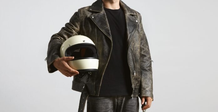 A person wearing a weathered gray leather motorcycle jacket, black t-shirt, and holding a cream-colored open-face helmet.