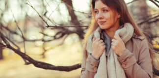 Travel Friendly Topcoat: Lightweight Style for Every Journey A woman standing outdoors in a fall setting, wearing a beige coat and a chunky gray scarf, holding the scarf near her neck and looking off to the side with a gentle smile. Blurred trees and warm tones create a cozy autumn mood. Travel Friendly Topcoat