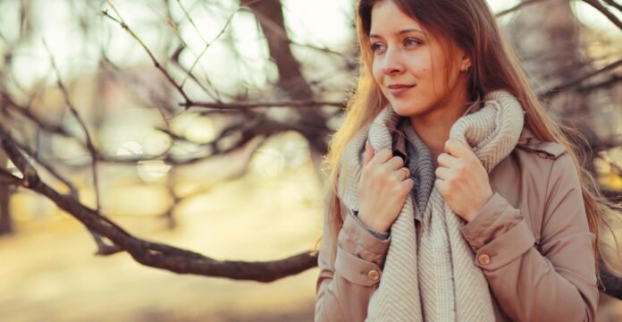 A woman standing outdoors in a fall setting, wearing a beige coat and a chunky gray scarf, holding the scarf near her neck and looking off to the side with a gentle smile. Blurred trees and warm tones create a cozy autumn mood. Travel Friendly Topcoat