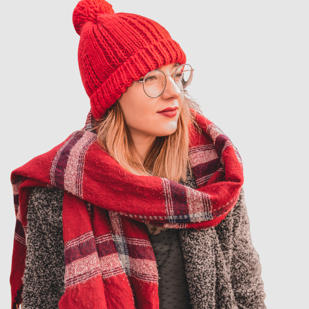 A woman wearing a bright red knit hat, round glasses, and a red plaid scarf wrapped around her shoulders against a light gray background.