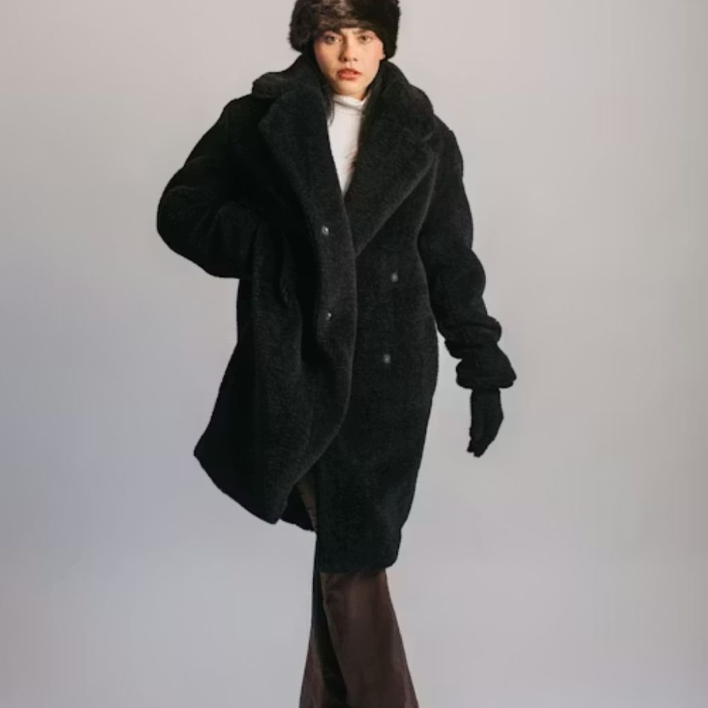 A person wearing a camel-colored belted coat stands with hands in pockets against a stone wall.Is a Long Parka Coat Worth It?