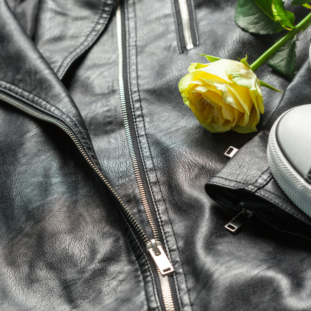 A close-up photo of a black zip-up leather jacket with a yellow rose tucked into the zipper area and a white sneaker toe visible at the edge.
