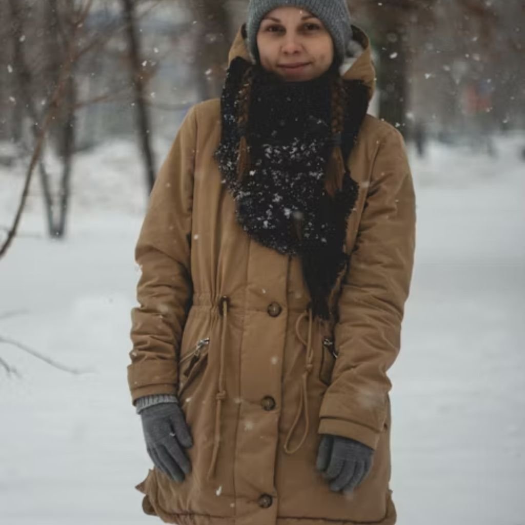 Person wearing a tan winter coat, gray knit hat, scarf, and gloves standing outdoors as snow falls in a park.Trench-Style Winter Coats