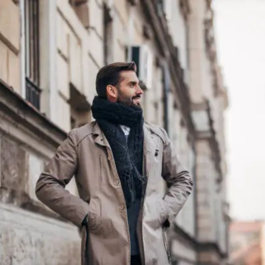 Man wearing a beige trench coat and black scarf standing on a city street, looking to the side.Trench-Style Winter Coats