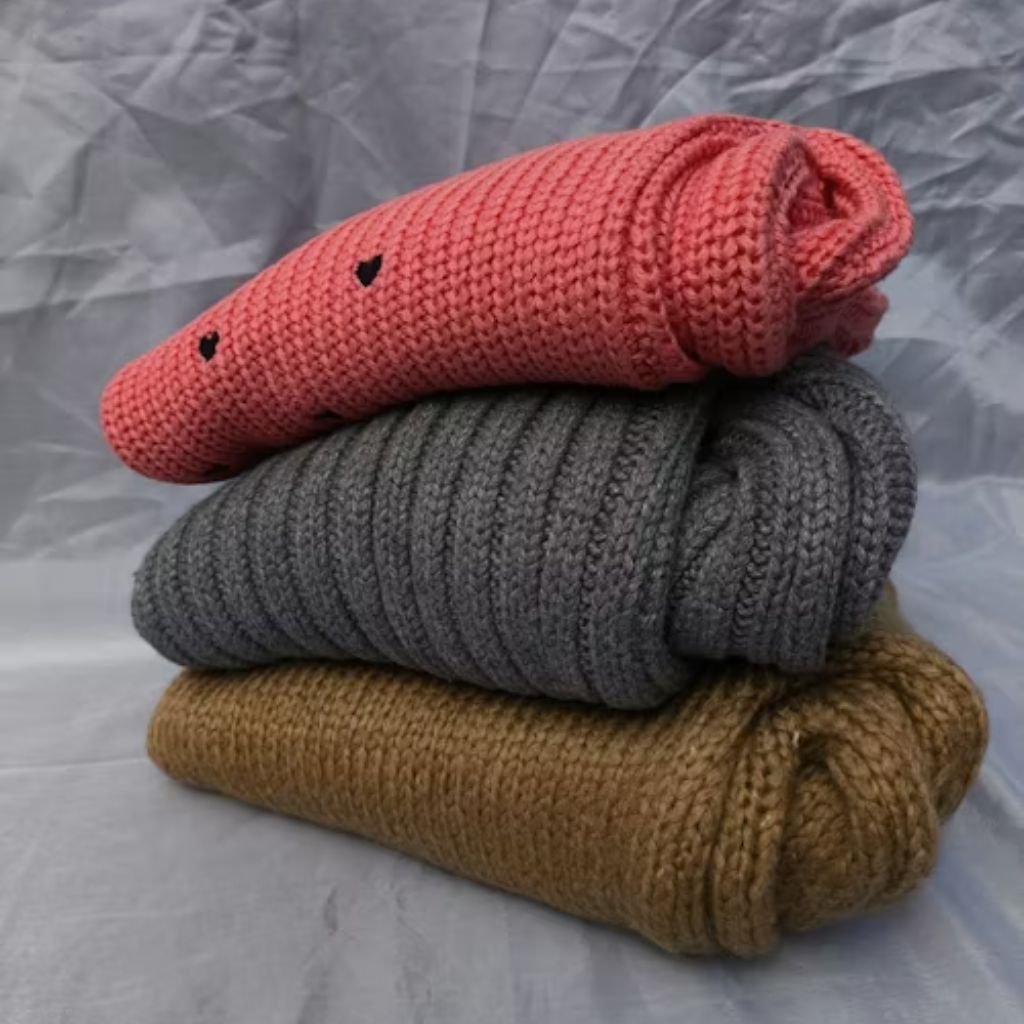 Stack of three folded knitted sweaters in pink, dark gray, and mustard yellow on a neutral fabric background.Is Herringbone Wool Worth It?