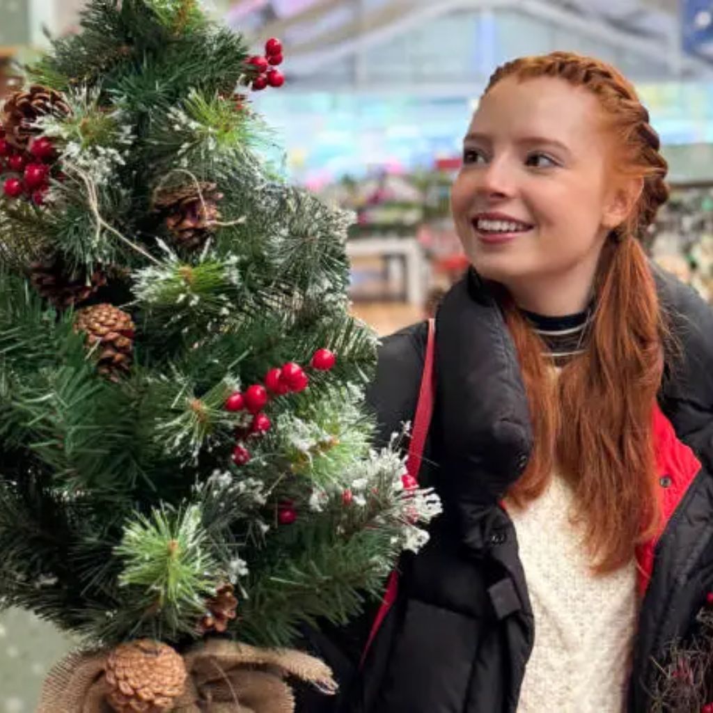 A smiling young woman with long red hair styled in braids looks at a decorated Christmas tree with berries and pinecones inside a festive indoor setting.Why Oversized Padded Jackets