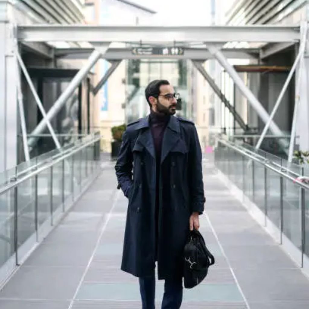 A man wearing a dark trench coat and glasses walks across a modern glass-covered walkway, holding a black leather bag and looking to the side.Trench-Style Winter Coats