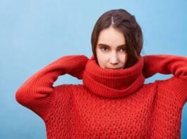 A woman wearing a bright red chunky-knit turtleneck sweater lifts her hands behind her head while standing against a light blue background.Why Mock neck turtlenecks