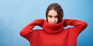 Why Mock Neck Turtlenecks Are Perfect for Cold-Weather Style A woman wearing a bright red chunky-knit turtleneck sweater lifts her hands behind her head while standing against a light blue background.Why Mock neck turtlenecks