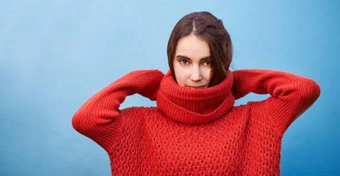 A woman wearing a bright red chunky-knit turtleneck sweater lifts her hands behind her head while standing against a light blue background.Why Mock neck turtlenecks