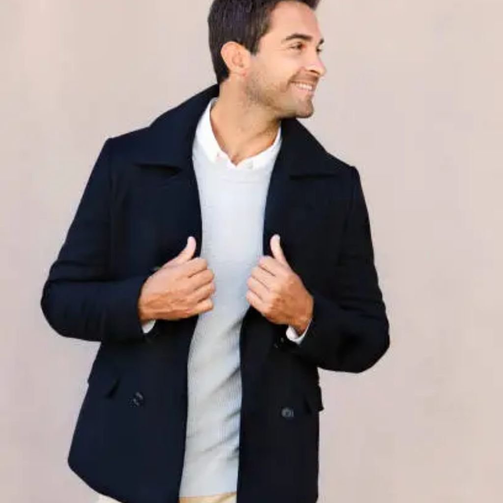 Person smiling and wearing a dark coat over a light sweater, looking to the side.The Wool Peacoat Comeback: