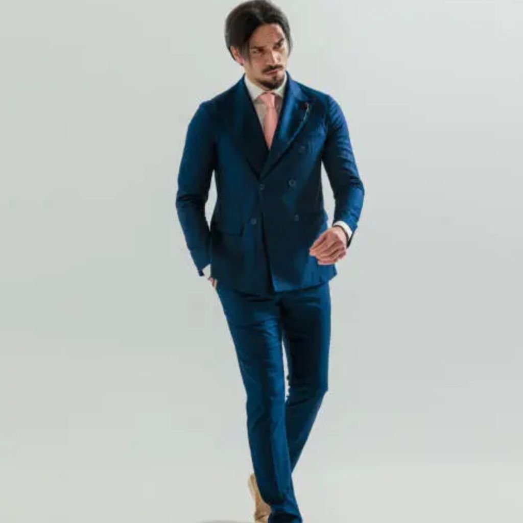 A man walking forward in a tailored blue double-breasted suit with a light pink tie and tan shoes against a plain light background.Double-Breasted Wool Jackets: