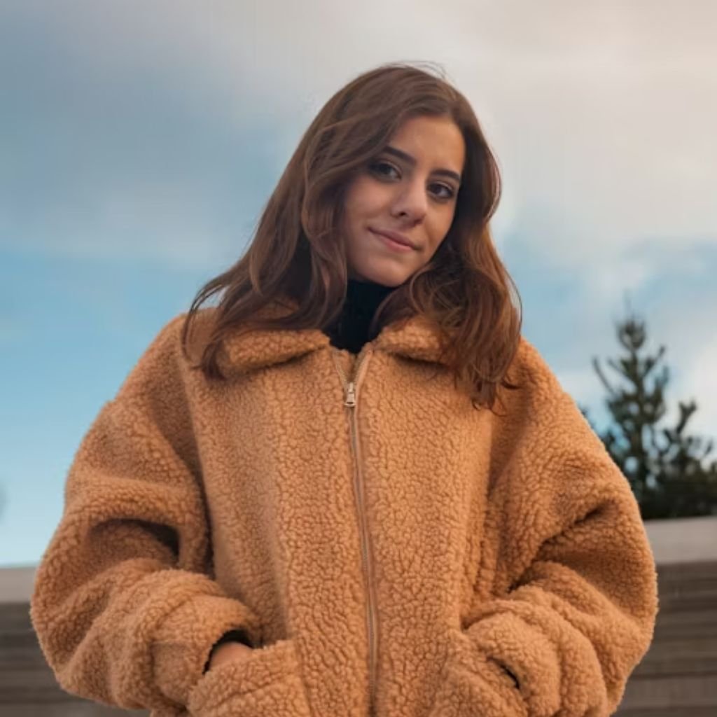 A person wearing a gray overcoat, black scarf, and sunglasses, standing against a dark background and adjusting their coat.The Wool Peacoat Comeback: