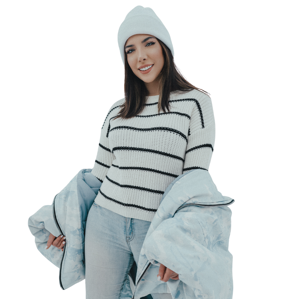 A young woman wearing a light gray beanie, a white knit sweater with black stripes, and light blue jeans. She holds a pale blue puffer jacket open and smiles at the camera against a plain white background.