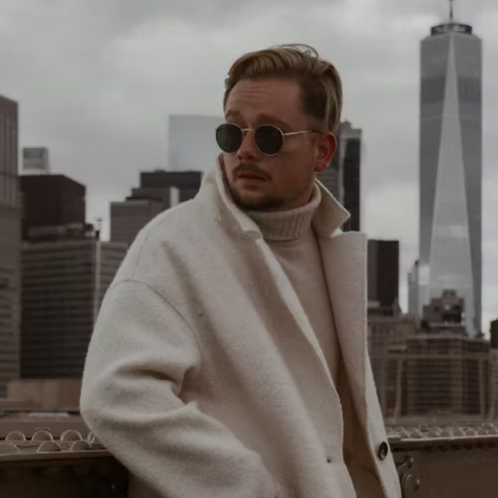 A person wearing round sunglasses and a light-colored coat stands on a bridge with a city skyline in the background.The Wool Peacoat Comeback:
