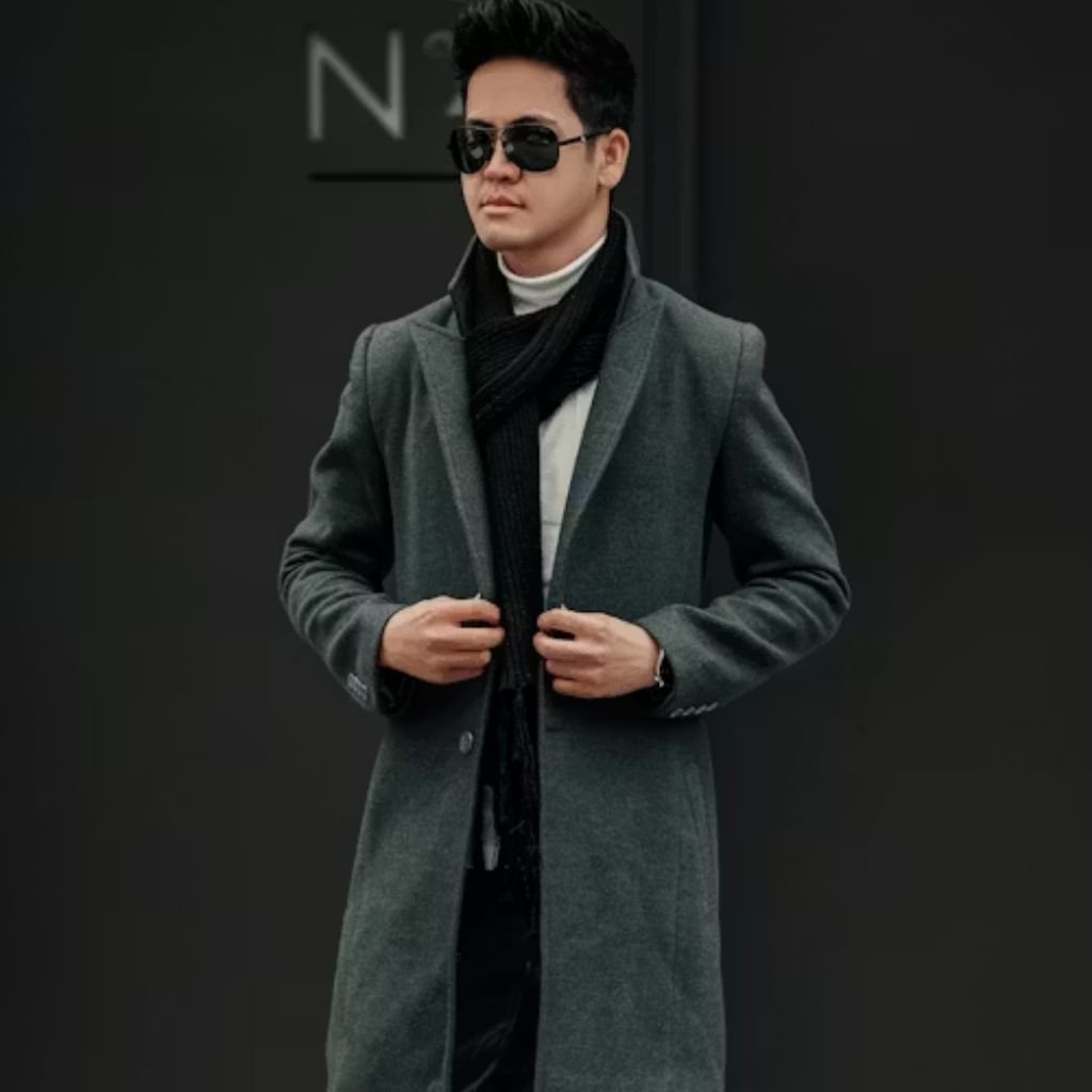 A person wearing a gray overcoat, black scarf, and sunglasses, standing against a dark background and adjusting their coat.The Wool Peacoat Comeback:
