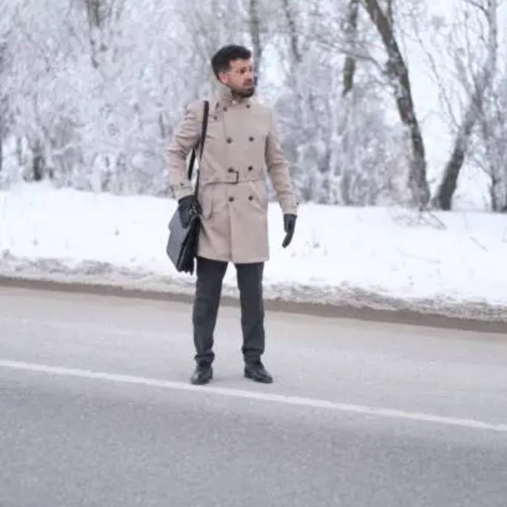 A bearded man wearing a tan suede coat with dark fur trim and a mustard turtleneck leans against a decorative black iron railing outdoors.Trench-Style Winter Coats