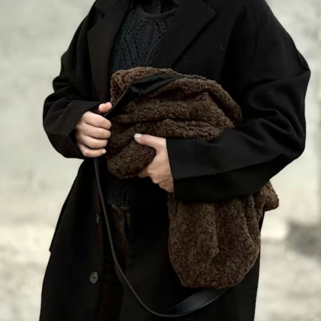 A man wearing a dark trench coat and glasses walks across a modern glass-covered walkway, holding a black leather bag and looking to the side.Trench-Style Winter Coats