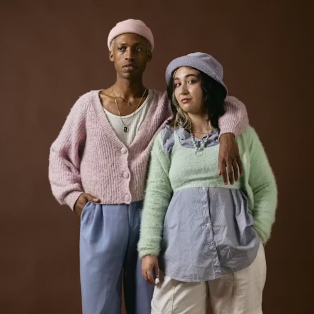 Two people standing side by side wearing pastel-colored knit sweaters and hats against a brown studio backdrop.Lambswool Clothing
