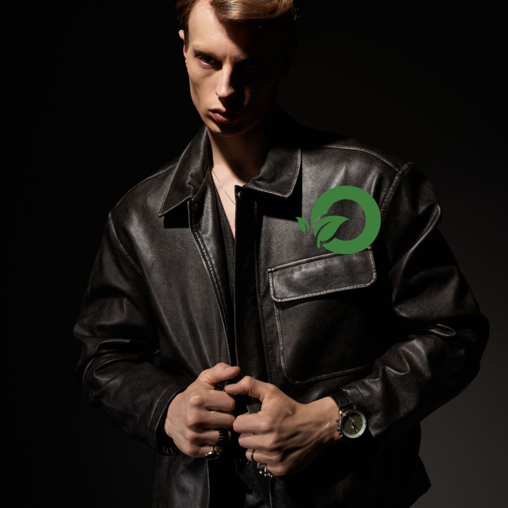 Workwear Field Leather Jacket: Durable Style for Everyday 5 A man wearing a black leather field jacket with a large front pocket zipper detail. He is posed against a dark background, with a green circular leaf emblem on the chest area.
