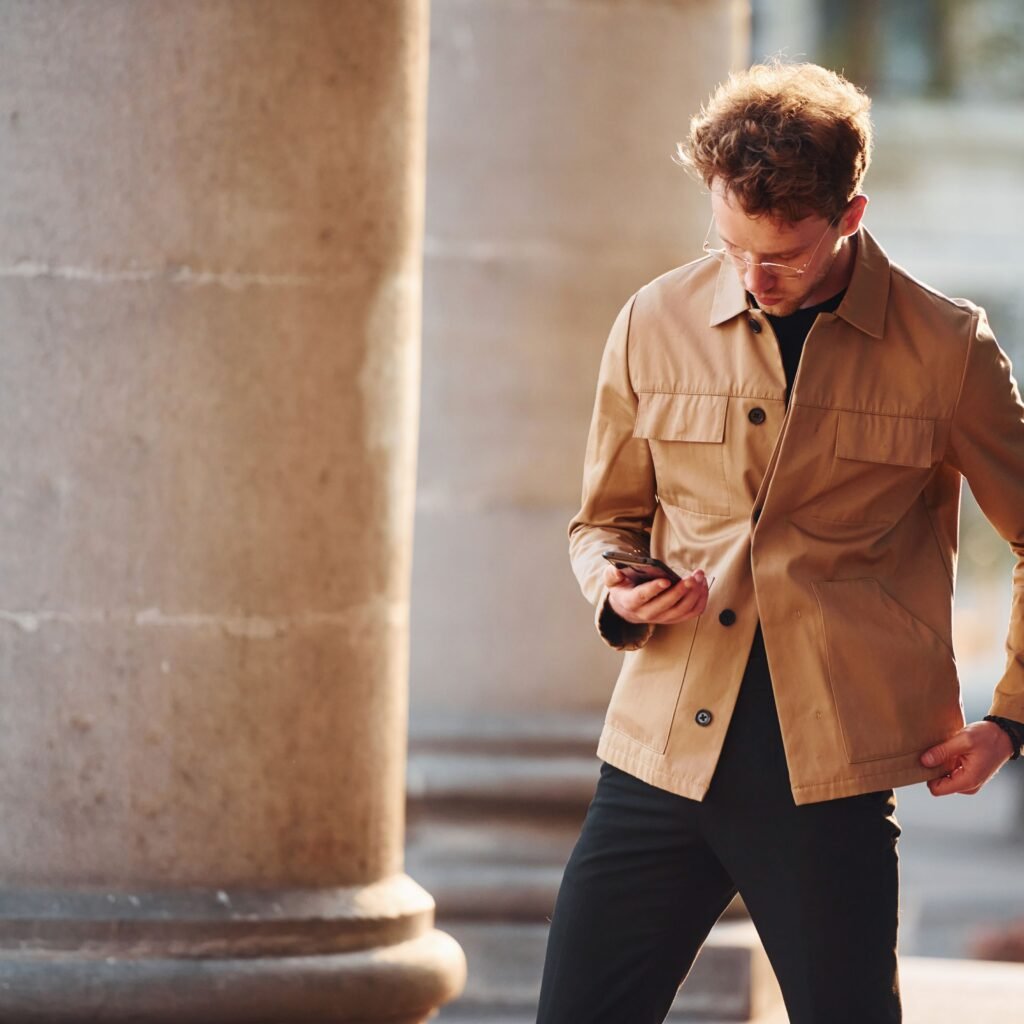 A man wearing a tan workwear field leather jacket, black pants, and glasses, walking outdoors near stone columns while looking at his phone.