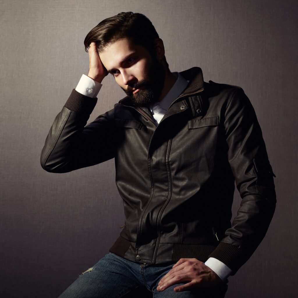 A man with a beard wearing a black leather travel-field jacket, white shirt cuffs visible, seated against a neutral backdrop.Travel Field Leather Jacket