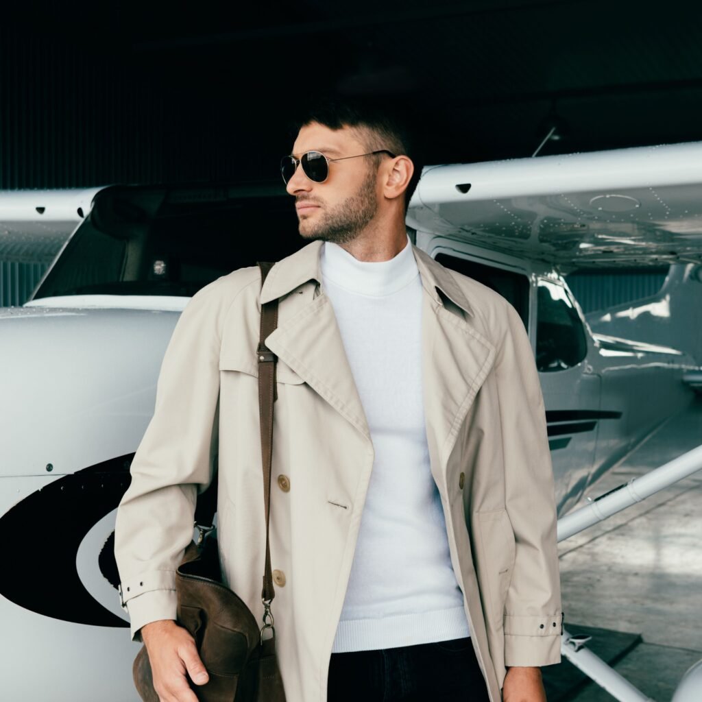A stylish man stands beside a small private airplane in a hangar. He wears a beige field leather jacket over a white knit turtleneck, dark jeans, and sunglasses, and carries a brown messenger bag slung over his shoulder.