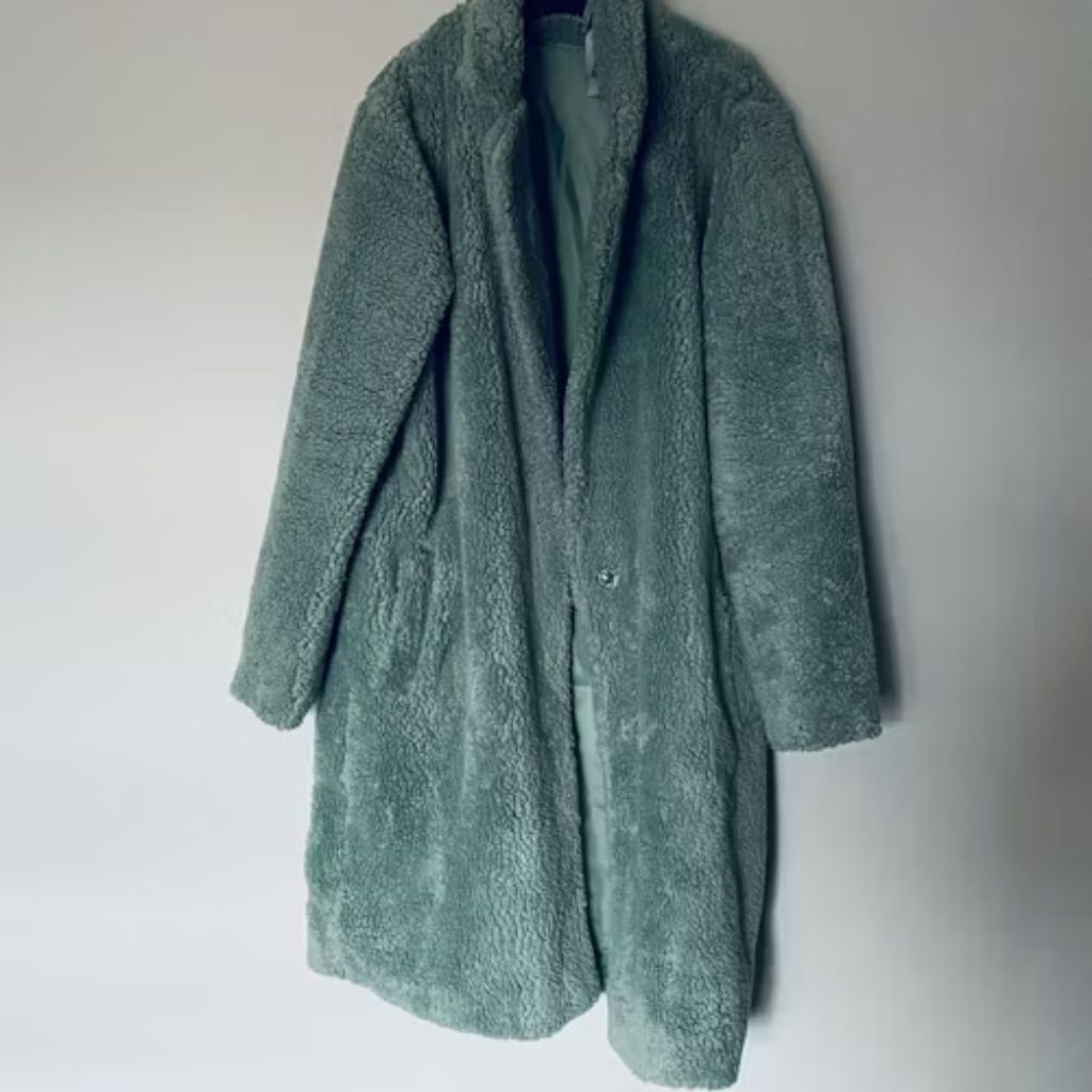 A long, sage-green teddy coat with a soft, textured fabric hanging against a plain wall.Boiled Wool Jacket