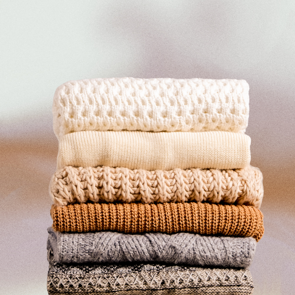 A stack of cozy knitted sweaters in neutral tones, arranged from light cream to dark beige and gray.