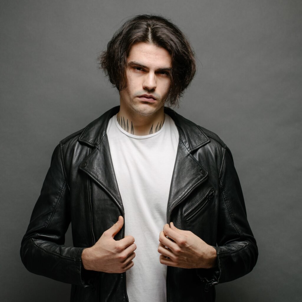  stylish young man with medium-length dark hair wearing a black leather biker jacket over a white t-shirt, gazing at the camera against a gray backdrop.