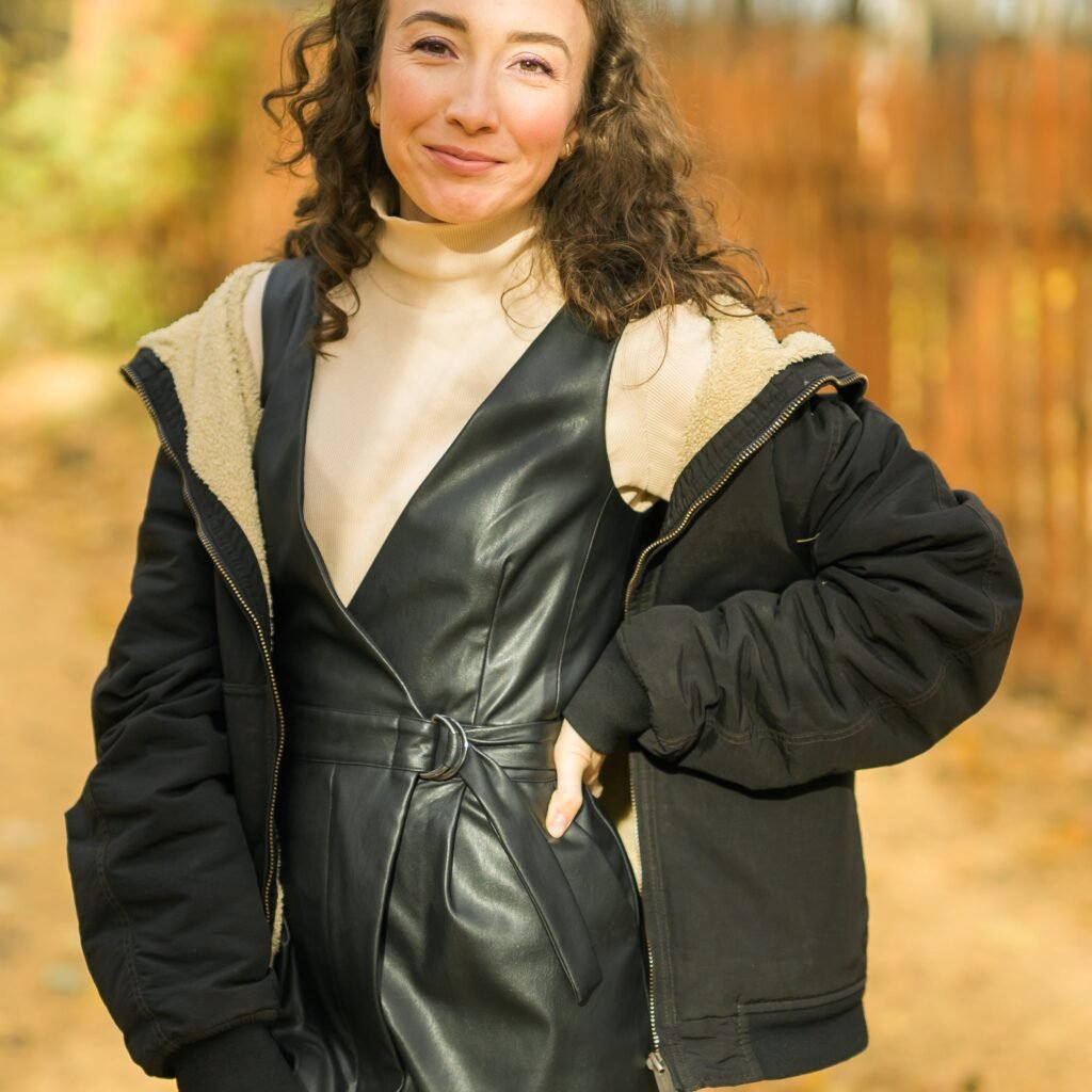 A stylish woman outdoors wearing a black leather belted jacket layered over a beige turtleneck, with a black oversized jacket over it. Autumnal background with warm lighting.
