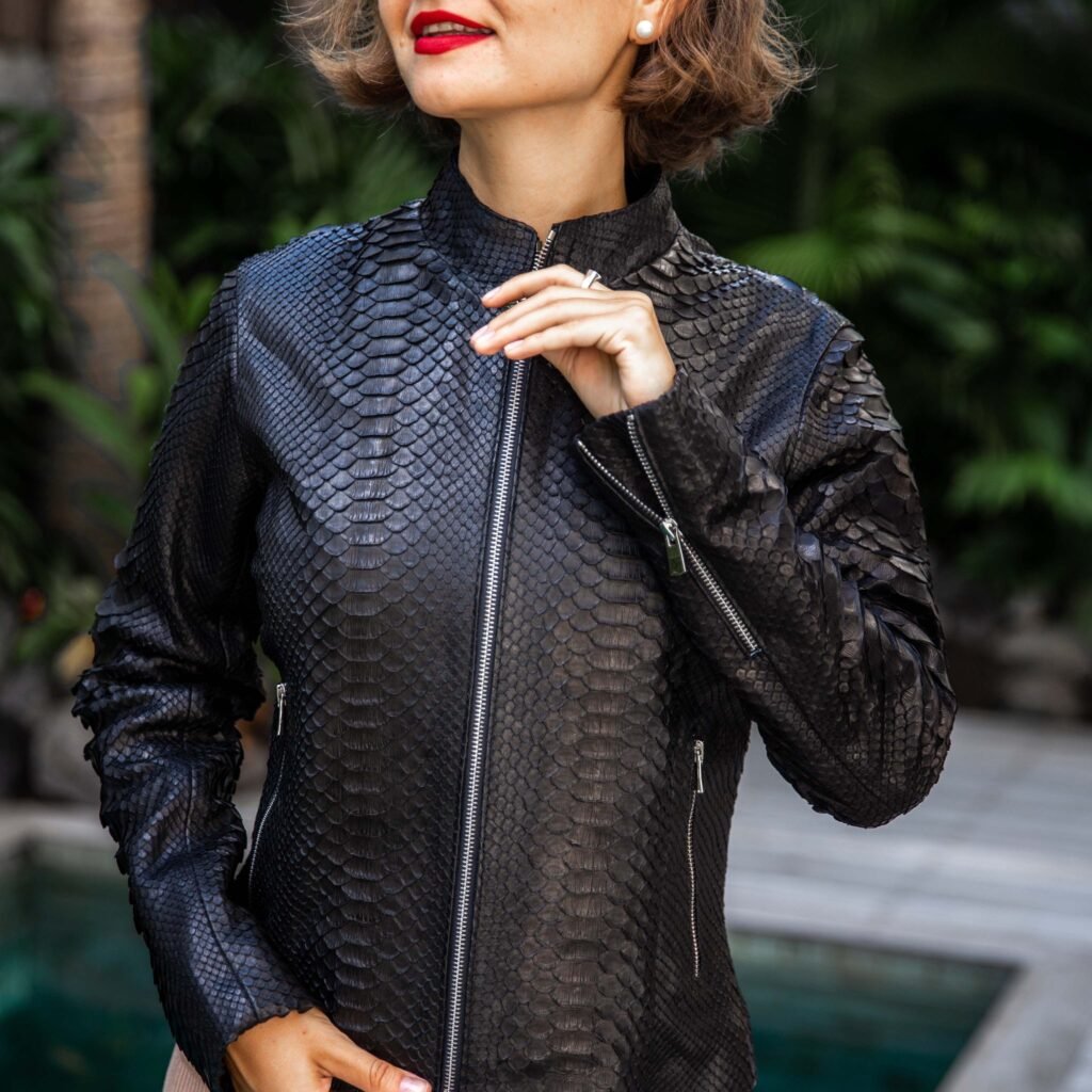 A stylish woman wearing a textured black leather jacket with a scale-like pattern, standing outdoors near greenery and a pool.

