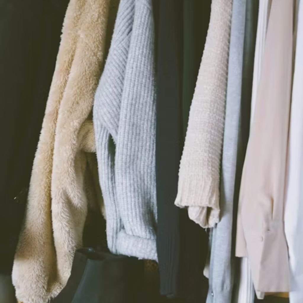 Neutral-toned sweaters and clothing hanging neatly on a wardrobe rack, showcasing soft knit and textured fabrics.The image shows a selection of clothing pieces hanging on a rack, primarily in soft, neutral shades such as beige, cream, grey, and off-white. The garments include cozy knit sweaters and textured fabrics, suggesting warmth and comfort, likely suited for cooler seasons. The simple color palette and natural materials create a calm, minimalist aesthetic, highlighting an organized wardrobe focused on timeless fashion and everyday versatility.
