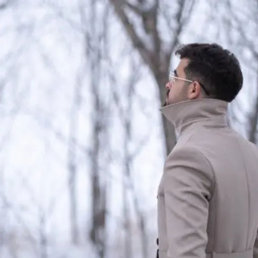 Person wearing a tan winter coat, gray knit hat, scarf, and gloves standing outdoors as snow falls in a park.Trench-Style Winter Coats