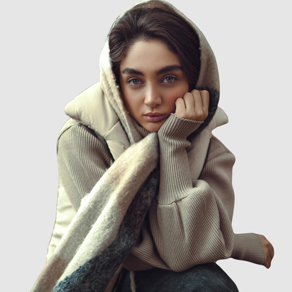 A young woman wearing a beige hoodie and cozy scarf, looking at the camera with her hand near her face.